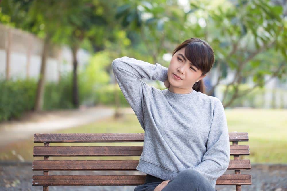 A woman sitting on a park bench holds her neck as she suffers from neck pain.